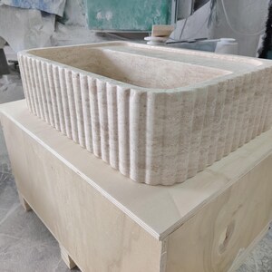 May include: A rectangular, beige stone sink with a fluted exterior design. The sink is placed on top of a light-colored wooden crate. The sink's interior is smooth, and the overall aesthetic is modern and minimalist.