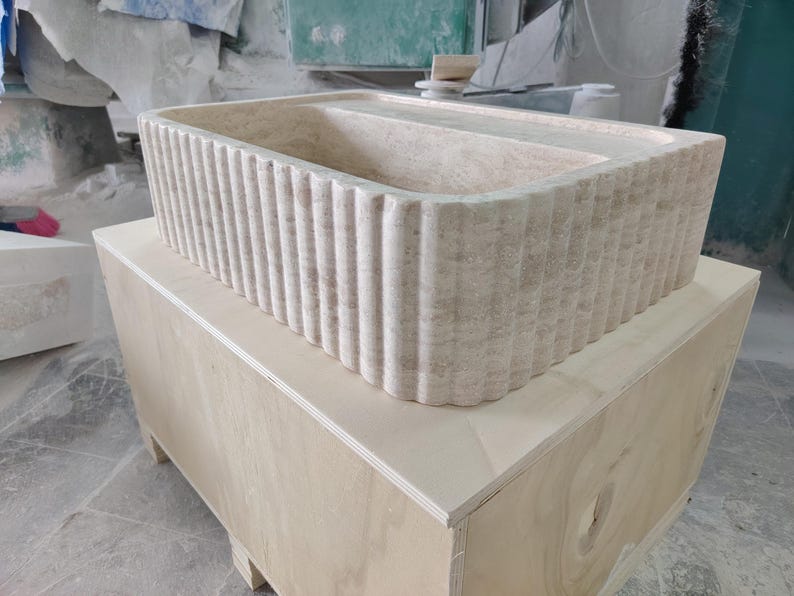 May include: A rectangular, beige stone sink with a fluted exterior design. The sink is placed on top of a light-colored wooden crate. The sink's interior is smooth, and the overall aesthetic is modern and minimalist.