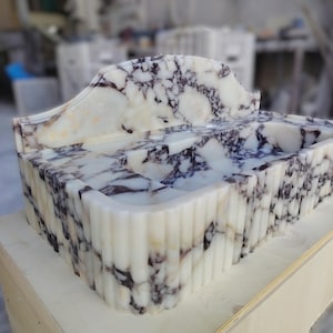 May include: A rectangular marble sink with a white and purple veined pattern. The sink has a curved backsplash and a fluted exterior. The sink is sitting on a wooden surface.