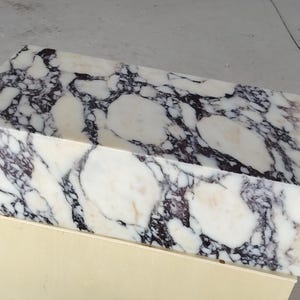 May include: A rectangular block of polished white marble featuring prominent, irregular dark purple and black veining throughout the surface. The stone displays a high-gloss finish with natural mineral patterns that create a bold, high-contrast appearance.