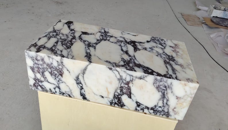 May include: A rectangular block of polished white marble featuring prominent, irregular dark purple and black veining throughout the surface. The stone displays a high-gloss finish with natural mineral patterns that create a bold, high-contrast appearance.