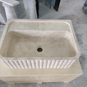 May include: A rectangular, beige stone sink with a fluted exterior design. The sink has a central drain hole and a raised back section. It is placed on a matching stone base, showcasing a natural, textured appearance.