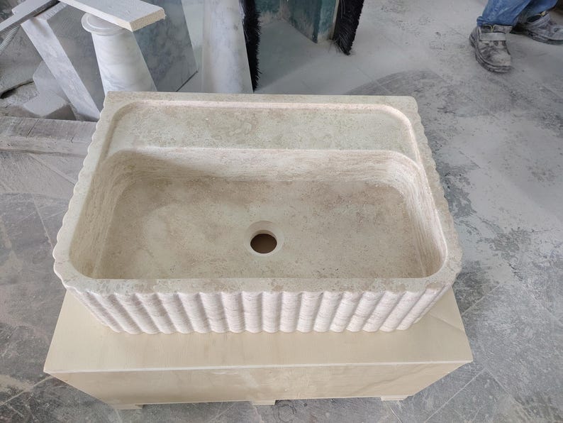 May include: A rectangular, beige stone sink with a fluted exterior design. The sink has a central drain hole and a raised back section. It is placed on a matching stone base, showcasing a natural, textured appearance.