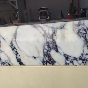 May include: A large rectangular block of polished white marble featuring prominent, irregular veining in shades of deep purple, black, and subtle golden-brown. The stone surface is smooth and reflective, showcasing a natural, complex pattern throughout.