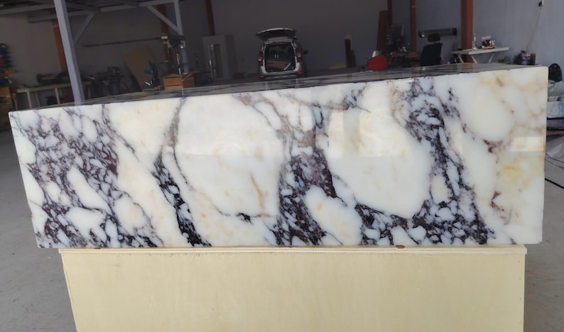 May include: A large rectangular block of polished white marble featuring prominent, irregular veining in shades of deep purple, black, and subtle golden-brown. The stone surface is smooth and reflective, showcasing a natural, complex pattern throughout.
