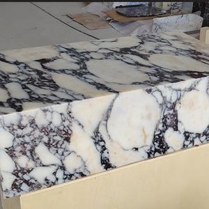 May include: A rectangular block of polished white marble featuring prominent, irregular black and dark purple veining throughout its surface. The stone block has sharp, clean edges and a smooth, reflective finish, showcasing natural mineral patterns.
