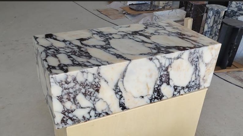 May include: A rectangular block of polished white marble featuring prominent, irregular black and dark purple veining throughout its surface. The stone block has sharp, clean edges and a smooth, reflective finish, showcasing natural mineral patterns.
