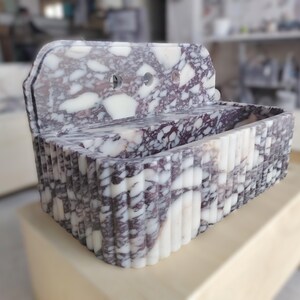 K&ouml;nnte beinhalten: Ein rechteckiges Waschbecken aus marmor&auml;hnlichem Material mit einer R&uuml;ckwand. Das Waschbecken weist ein Muster aus wei&szlig;en und dunkelvioletten Adern auf. Die Vorderseite des Waschbeckens hat eine geriffelte Textur. Zwei runde L&ouml;cher sind in der R&uuml;ckwand zu sehen.