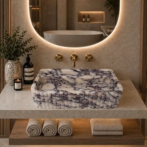 May include: A rectangular marble sink with a purple and white pattern sits on a light-colored countertop. The sink is the focal point of a modern bathroom, with gold fixtures and a round mirror. Rolled towels and a candle are visible.