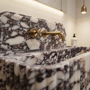 K&ouml;nnte beinhalten: Ein Waschbecken aus Marmor mit einem dunkelvioletten und wei&szlig;en Muster. Das Waschbecken hat einen goldenen Wasserhahn und Armaturen. Das Waschbecken ist rechteckig mit einem geriffelten Design auf der Vorderseite. Eine goldene Leuchte h&auml;ngt dar&uuml;ber.