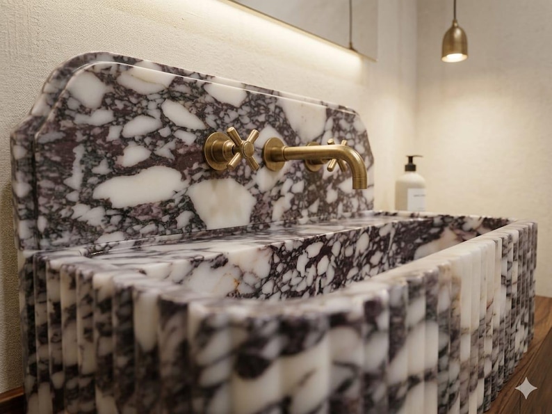 K&ouml;nnte beinhalten: Ein Waschbecken aus Marmor mit einem dunkelvioletten und wei&szlig;en Muster. Das Waschbecken hat einen goldenen Wasserhahn und Armaturen. Das Waschbecken ist rechteckig mit einem geriffelten Design auf der Vorderseite. Eine goldene Leuchte h&auml;ngt dar&uuml;ber.