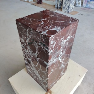 May include: A rectangular pedestal made of polished deep red marble with prominent white veining throughout its surface. The stone block features sharp, clean edges and a smooth, reflective finish, showcasing the natural patterns of the rock.