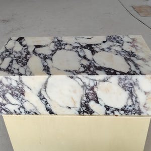 May include: A rectangular block of polished white marble featuring prominent, irregular veining in shades of deep purple, black, and soft cream. The stone surface displays a high-gloss finish with a complex, organic pattern of interlocking shapes.