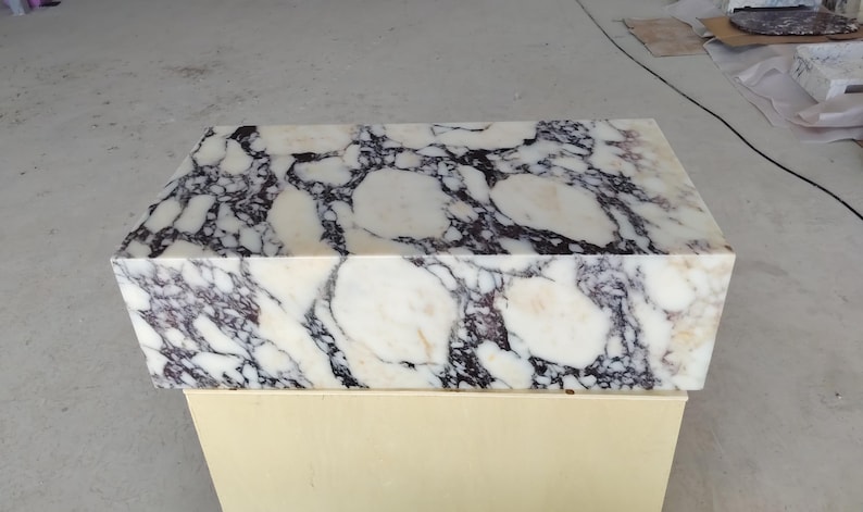 May include: A rectangular block of polished white marble featuring prominent, irregular veining in shades of deep purple, black, and soft cream. The stone surface displays a high-gloss finish with a complex, organic pattern of interlocking shapes.