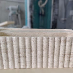 May include: A rectangular, off-white stone planter with vertical ribbed detailing. The planter has a simple, modern design and is suitable for indoor or outdoor use. The dimensions are approximately 24 inches long, 10 inches wide, and 8 inches high.