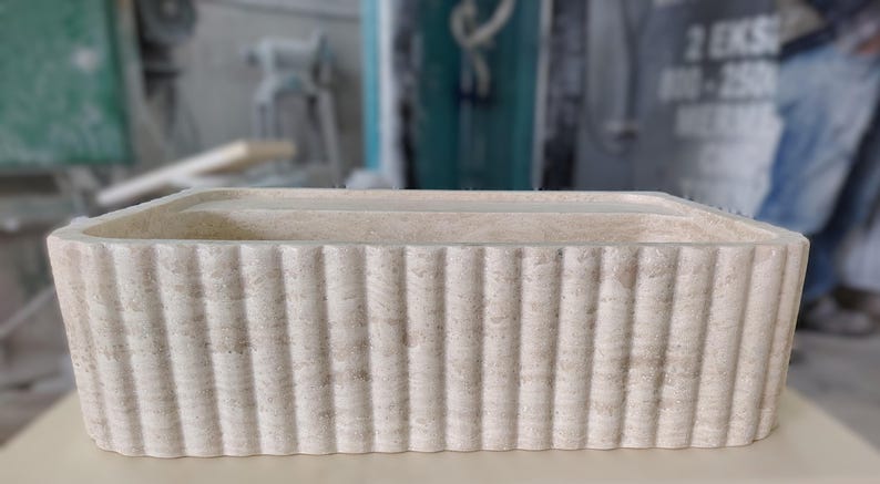 May include: A rectangular, off-white stone planter with vertical ribbed detailing. The planter has a simple, modern design and is suitable for indoor or outdoor use. The dimensions are approximately 24 inches long, 10 inches wide, and 8 inches high.