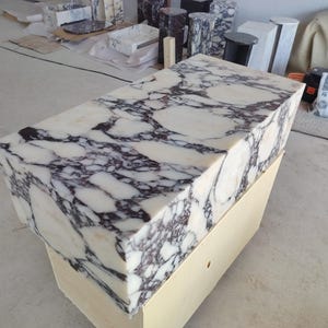 May include: A rectangular block of white marble featuring prominent, dark purple and black veining throughout its surface. The stone block is positioned atop a plain, light-colored wooden base, showcasing a polished finish on the top and sides.