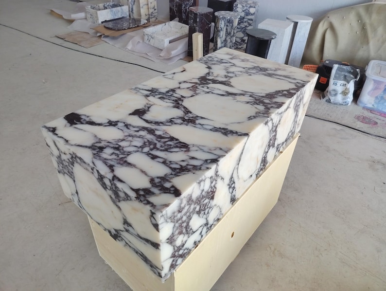 May include: A rectangular block of white marble featuring prominent, dark purple and black veining throughout its surface. The stone block is positioned atop a plain, light-colored wooden base, showcasing a polished finish on the top and sides.