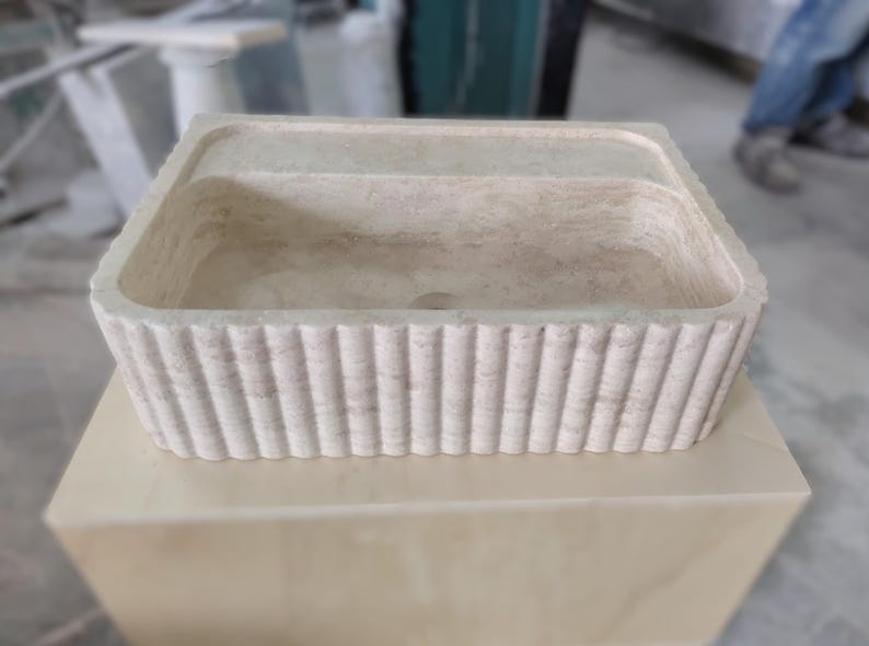May include: A rectangular, off-white stone sink with a fluted exterior. The sink is set on a light-colored, square base. The design features a central basin and a raised rim, suitable for bathroom or kitchen use.