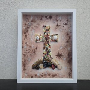 Könnte beinhalten: Gerahmtes Kunstwerk mit einem Kreuzdesign. Das Kreuz besteht aus verschiedenen bunten Steinen und Kieselsteinen, vor einem hellbraunen Hintergrund mit sternförmigen Mustern. Das Kunstwerk ist in einem weißen Rahmen eingefasst.