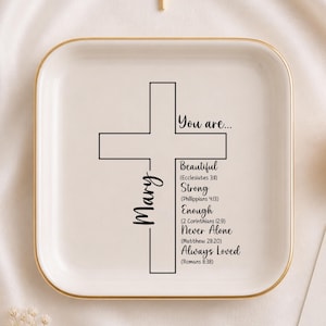 Puede incluir: Una bandeja de cerámica blanca con borde dorado presenta un diseño de cruz negra y las palabras "You are..." seguidas de frases inspiradoras. También hay una pequeña cruz dorada y una vela encendida.