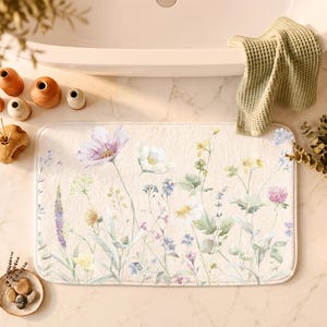 May include: A rectangular bath mat with a cream-colored base featuring a watercolor-style print of various wildflowers in shades of purple, blue, yellow, and white, with green stems and leaves scattered across the surface.