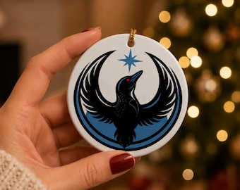 May include: A round white ceramic ornament featuring a black bird with outstretched wings, a blue outline, and a blue star. The bird has a red eye. A gold string is attached for hanging. The ornament is a holiday decoration.