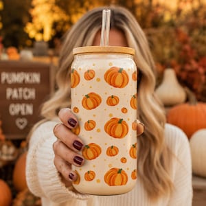 May include: A clear glass tumbler with a bamboo lid and a glass straw, featuring a repeating pattern of orange pumpkins and small yellow dots on a light cream background. The cylindrical glass is held upright, showcasing the festive autumn design.