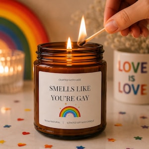 Peut inclure: Une bougie en pot de verre brun allumée avec une étiquette blanche qui dit "Smells Like You're Gay" avec un arc-en-ciel. Une allumette allume la bougie. Un arc-en-ciel et une tasse avec "Love is Love" sont en arrière-plan.