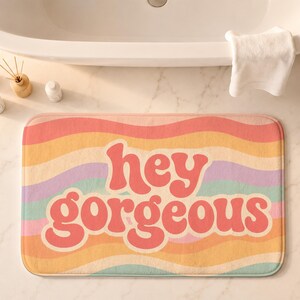 Puede incluir: Una alfombrilla de ba&ntilde;o rectangular con un patr&oacute;n de rayas onduladas multicolores en tonos rosa, naranja, amarillo, morado y turquesa. El texto "hey gorgeous" est&aacute; impreso en letras rosas grandes, negritas y redondeadas en el centro.
