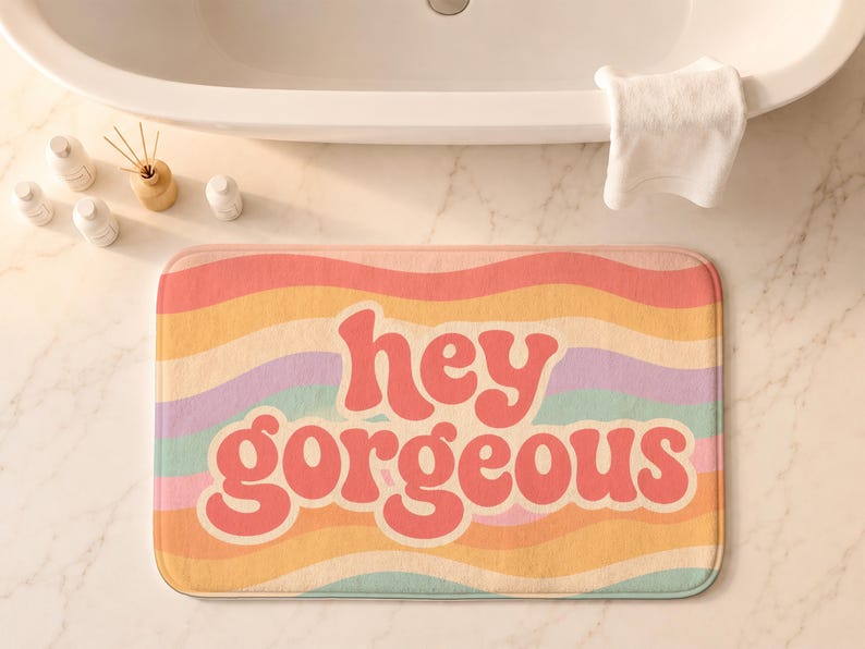 Puede incluir: Una alfombrilla de ba&ntilde;o rectangular con un patr&oacute;n de rayas onduladas multicolores en tonos rosa, naranja, amarillo, morado y turquesa. El texto "hey gorgeous" est&aacute; impreso en letras rosas grandes, negritas y redondeadas en el centro.