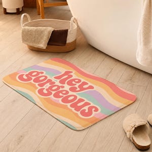 Puede incluir: Una alfombrilla de ba&ntilde;o rectangular con un patr&oacute;n de rayas onduladas multicolores en tonos rosa, naranja, amarillo, morado y turquesa. El texto "hey gorgeous" est&aacute; impreso en una fuente rosa oscuro, redondeada y negrita en el centro.