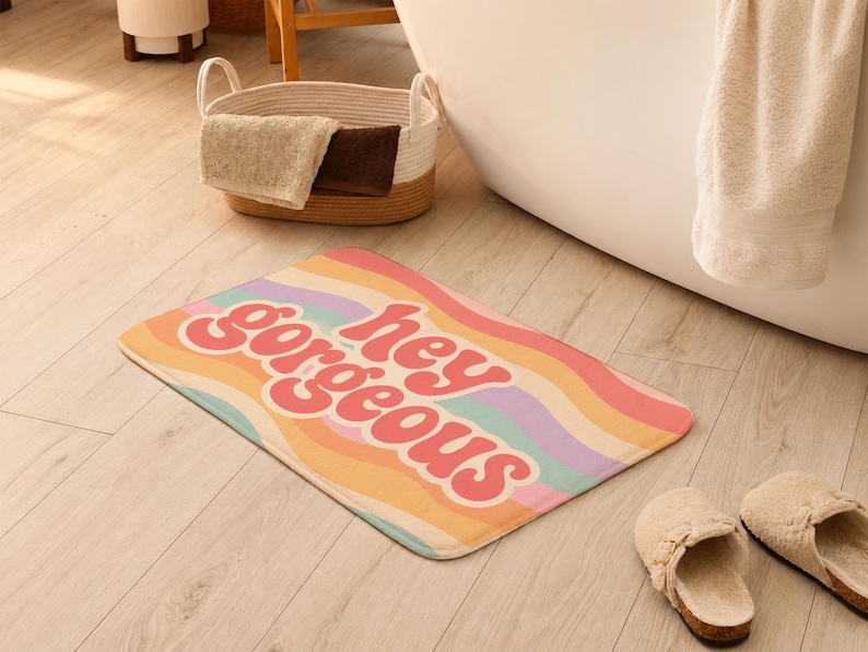 Puede incluir: Una alfombrilla de ba&ntilde;o rectangular con un patr&oacute;n de rayas onduladas multicolores en tonos rosa, naranja, amarillo, morado y turquesa. El texto "hey gorgeous" est&aacute; impreso en una fuente rosa oscuro, redondeada y negrita en el centro.
