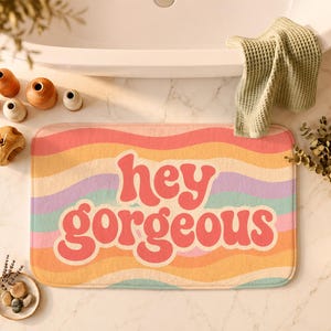 Puede incluir: Una alfombrilla de ba&ntilde;o rectangular con un patr&oacute;n de rayas onduladas multicolores en tonos rosa, naranja, amarillo, morado y turquesa. El texto "hey gorgeous" est&aacute; impreso en letras rojas grandes, negritas y redondeadas.