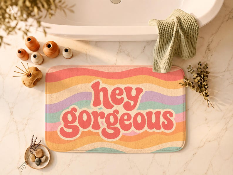 Puede incluir: Una alfombrilla de ba&ntilde;o rectangular con un patr&oacute;n de rayas onduladas multicolores en tonos rosa, naranja, amarillo, morado y turquesa. El texto "hey gorgeous" est&aacute; impreso en letras rojas grandes, negritas y redondeadas.
