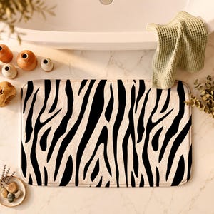 May include: A rectangular bath mat featuring a high-contrast black and white zebra stripe pattern. The mat has rounded corners and a soft, plush texture, providing a bold graphic element that covers the surface with repeating animal print lines.