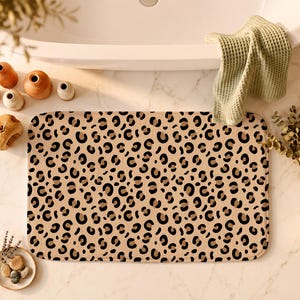 May include: A rectangular bath mat featuring a repeating leopard print pattern in shades of black, dark brown, and tan. The mat has rounded corners and a soft, textured surface, providing a functional floor covering for a bathroom space.