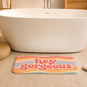 Puede incluir: Una alfombrilla de ba&ntilde;o rectangular con un patr&oacute;n de rayas onduladas multicolores en tonos rosa, naranja, amarillo, morado y verde azulado. El texto "hey gorgeous" est&aacute; impreso en una fuente rosa oscuro, negrita y redondeada en el centro.
