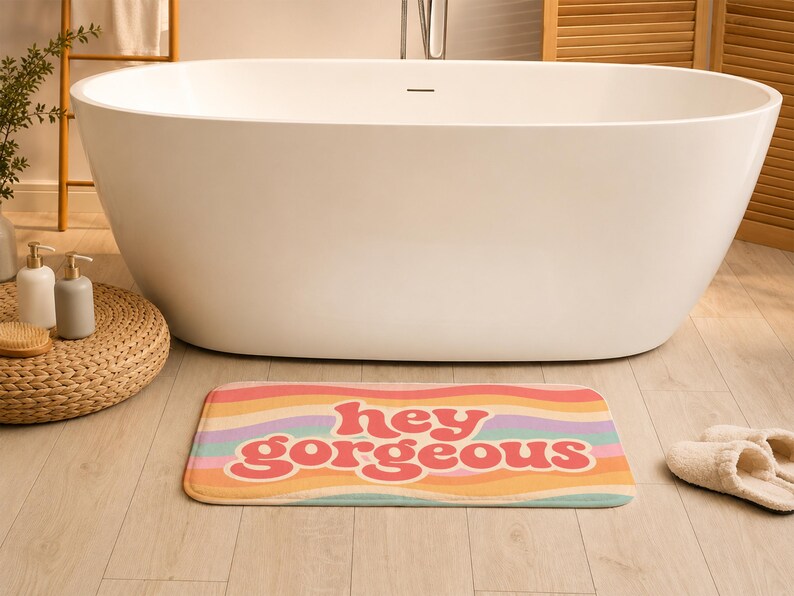 Puede incluir: Una alfombrilla de ba&ntilde;o rectangular con un patr&oacute;n de rayas onduladas multicolores en tonos rosa, naranja, amarillo, morado y verde azulado. El texto "hey gorgeous" est&aacute; impreso en una fuente rosa oscuro, negrita y redondeada en el centro.
