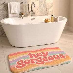 Puede incluir: Una alfombrilla de ba&ntilde;o rectangular con un patr&oacute;n de rayas onduladas multicolores en tonos rosa, naranja, amarillo y azul claro. El texto "hey gorgeous" est&aacute; impreso en letras rosas grandes, negritas y redondeadas en el centro.