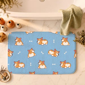 May include: A rectangular bath mat featuring a light blue background with a repeating pattern of cartoon Corgi dogs in various poses and small white bone shapes scattered throughout the design, creating a playful and consistent graphic motif.