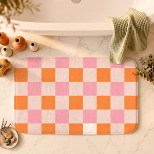 Puede incluir: Una alfombrilla de baño rectangular con un patrón de cuadros repetitivo en tonos rosa suave y naranja vibrante. La alfombrilla tiene esquinas redondeadas y una superficie texturizada y afelpada, con un diseño de cuadrícula uniforme.