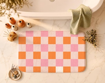 Puede incluir: Una alfombrilla de baño rectangular con un patrón de cuadros repetitivo en tonos rosa suave y naranja vibrante. La alfombrilla tiene esquinas redondeadas y una superficie texturizada y afelpada, con un diseño de cuadrícula uniforme.
