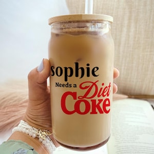 May include: A clear glass tumbler filled with a light brown beverage, ice cubes, and a straw. The glass has a wooden lid and features the text "sophie Needs a Diet Coke" in black and red lettering.