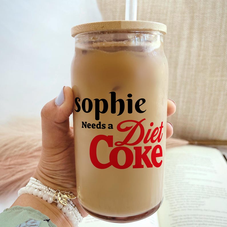 May include: A clear glass tumbler filled with a light brown beverage, ice cubes, and a straw. The glass has a wooden lid and features the text "sophie Needs a Diet Coke" in black and red lettering.