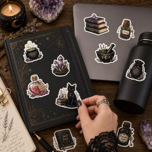 May include: A collection of mystical-themed stickers featuring a black cat, a cauldron, a stack of books, crystal clusters, potion bottles, and a pouch with a pentacle. The stickers are displayed on a black notebook and a dark gray laptop.