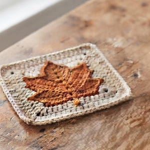 May include: A square crochet coaster featuring a central rust-coloured maple leaf motif surrounded by a light beige border. The coaster is constructed with a textured stitch pattern and has a flat, square shape with visible yarn loops.