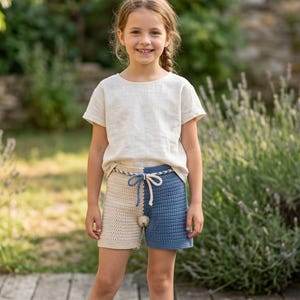 May include: A young girl wearing a cream-colored short-sleeved shirt and crochet shorts. The shorts are split into two colors, cream and blue, with a drawstring waist. The girl is standing on a wooden deck in front of a garden.