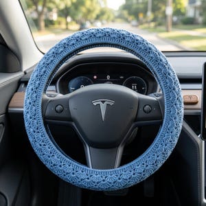 以下が含まれることがあります： 水色の、かぎ針編みのステアリングホイールカバー。カバーは、テクスチャ加工されたオープンウィーブパターンで、テスラのロゴが付いた黒いステアリングホイールに巻き付けられています。車のインテリアが背景に見えます。