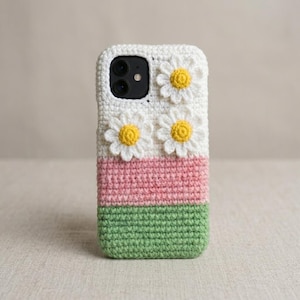 May include: A white, pink, and green crochet phone case with three-dimensional daisy accents. The case is divided into three horizontal sections: white at the top, pink in the middle, and green at the bottom. The daisies have white petals and yellow centers.